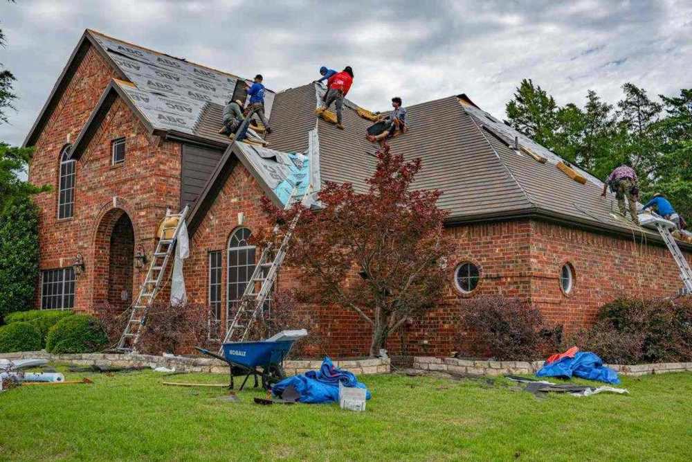A roofing crew replacing shingles on a brick house in Allen, Texas, with tarps and tear-off debris on the lawn below