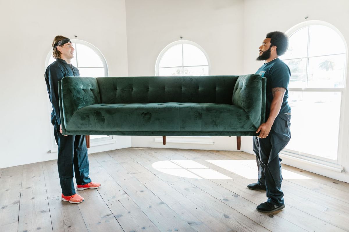 Two workers carrying a green sofa across an empty room