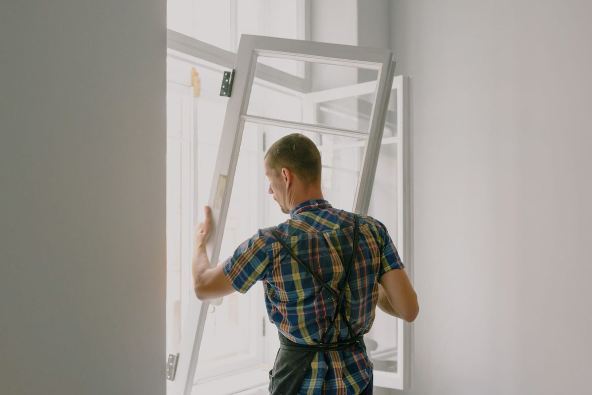 A worker in a plaid shirt lifting an old white window sash out of its frame