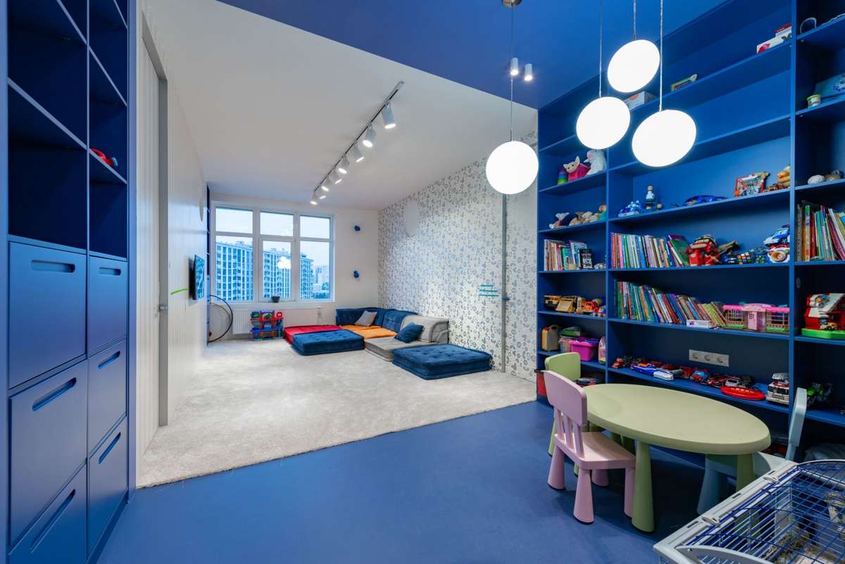 A children's playroom with blue shelves of books and toys, a small table and chairs, and floor cushions